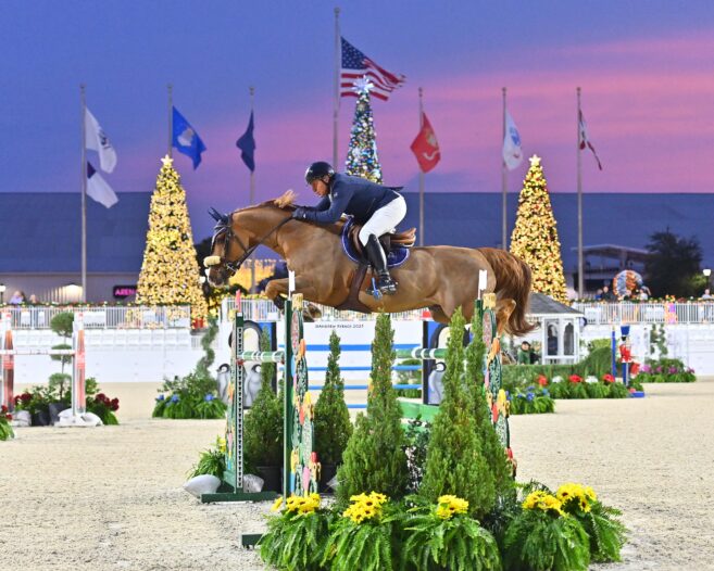 An equestrian rider and horse jump over an obstacle at an outdoor arena decorated with flags and lit Christmas trees during sunset.