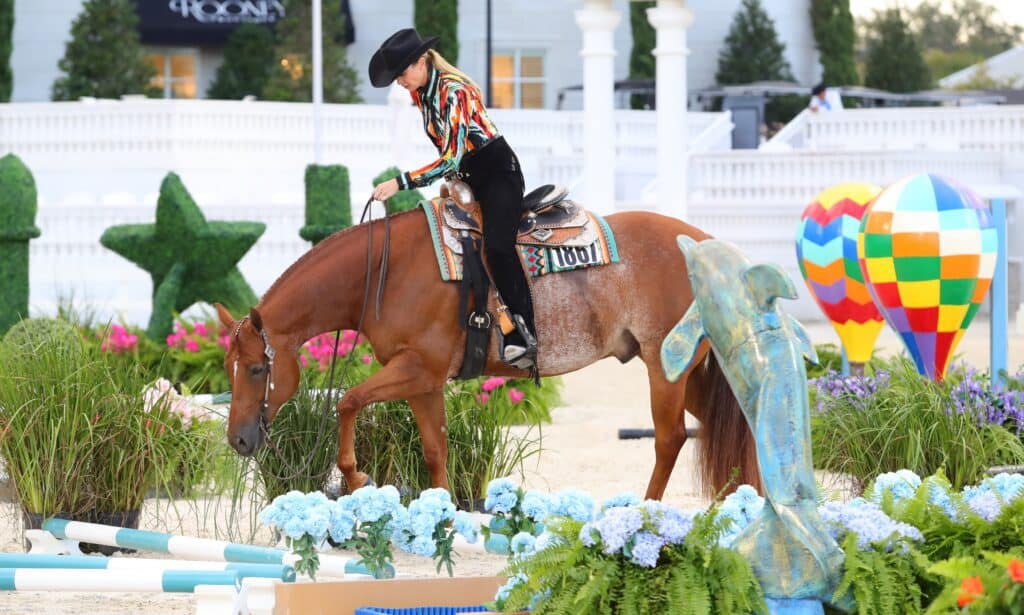 A person in a colorful shirt and black hat rides a brown horse through a decorated obstacle course with flowers and statues.