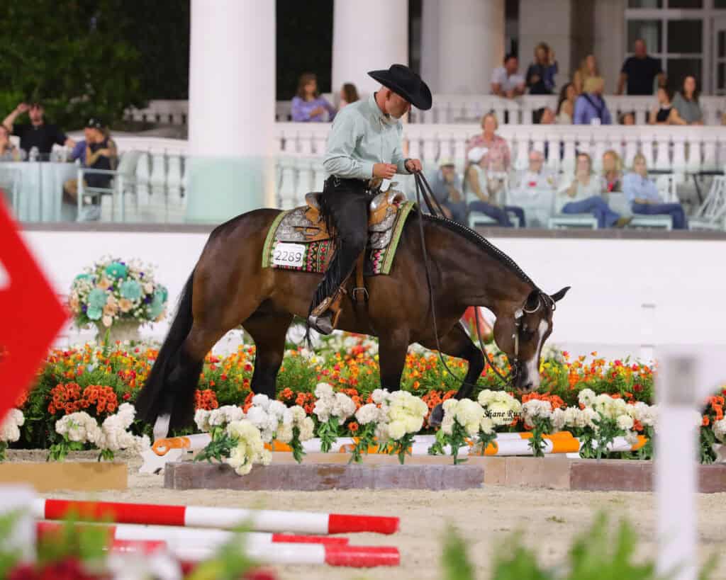 Man in a black cowboy hat rides a brown horse through an arena decorated with colorful flowers, with spectators seated in the background.