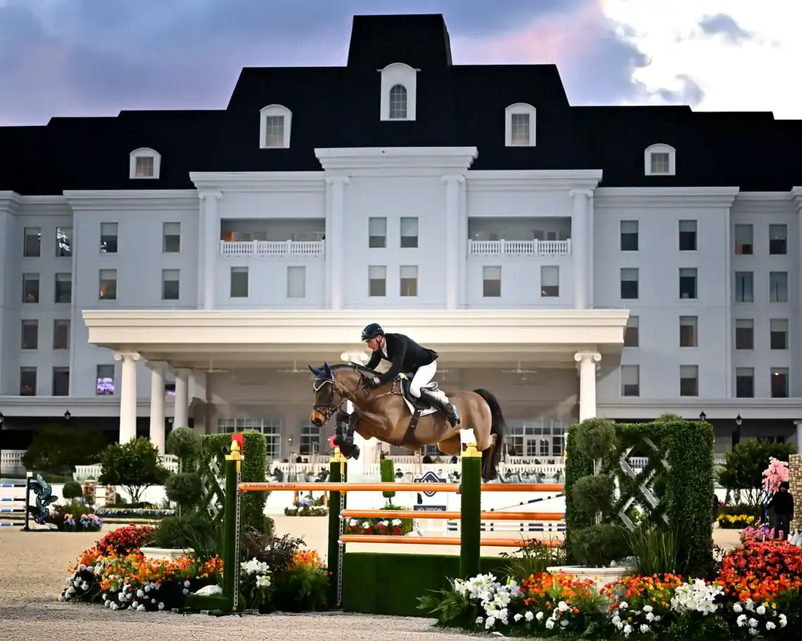 A horse and rider jump over an obstacle during an equestrian event in front of a large, white building with black roof and columns.
