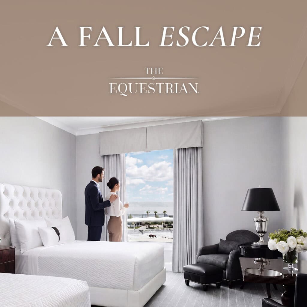 A man and woman stand by a window in a hotel room with a view of an equestrian event outside. Room features two beds, a black armchair, a lamp, and neutral decor.