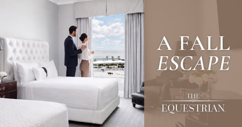 A couple stands by a window in a modern hotel room with two beds, overlooking a racetrack. Text reads: “A Fall Escape. The Equestrian.”.