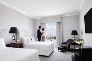 A couple stands by a window in a modern hotel room with two beds, black furniture, and a view of the ocean and palm trees outside.