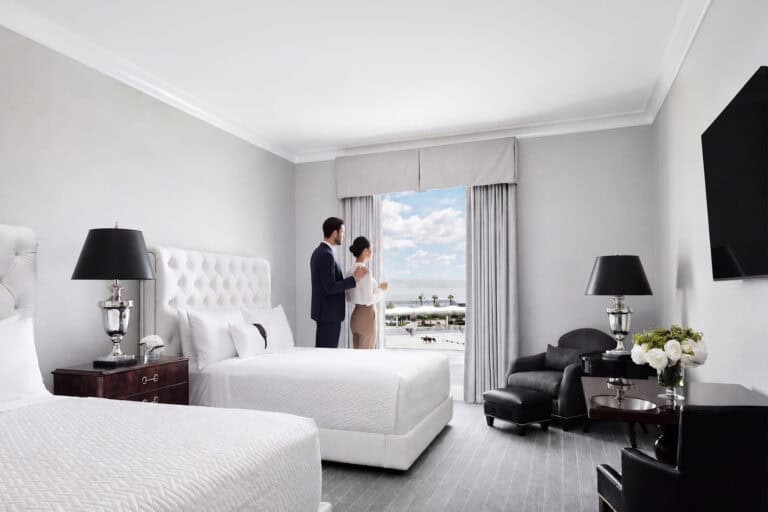 A couple stands by a window in a modern hotel room with two beds, black furniture, and a view of the ocean and palm trees outside.