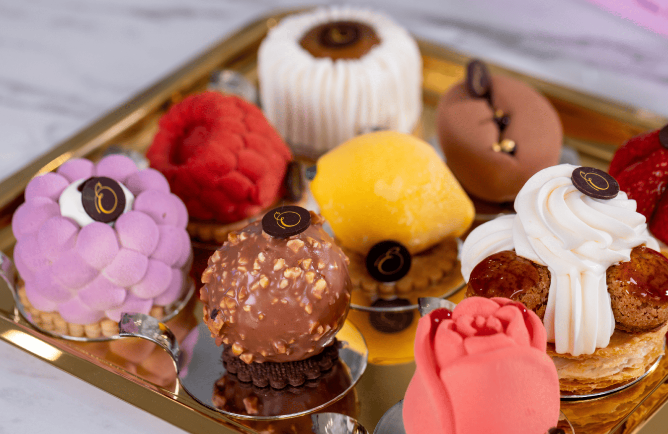 A gold tray holds assorted colorful, elaborately decorated pastries, including spheres, a rose-shaped dessert, and confections topped with chocolate emblems.