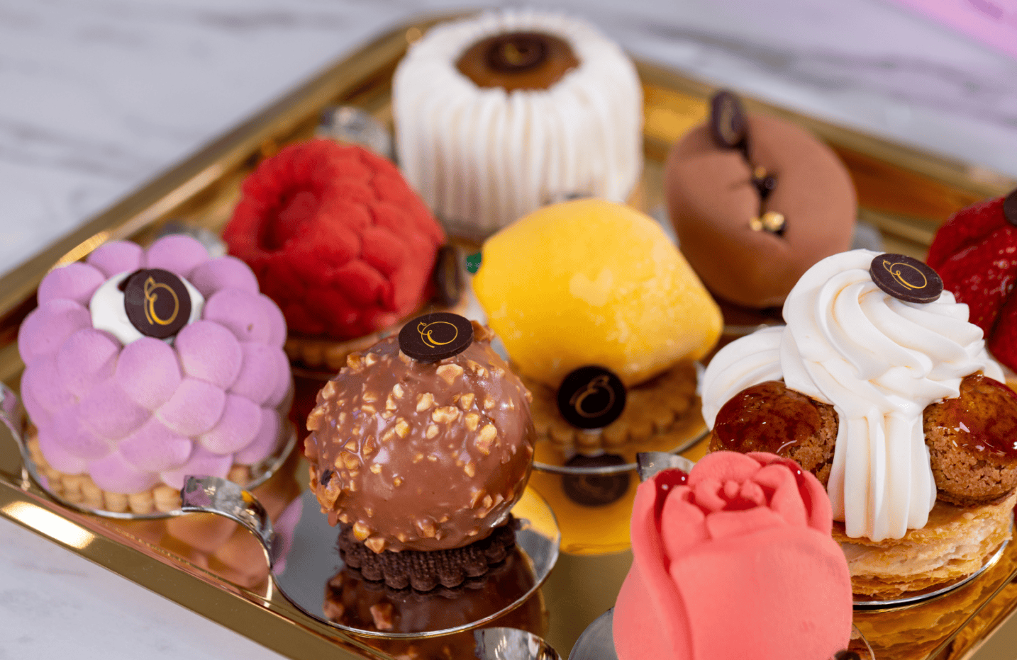 A gold tray holds assorted colorful, elaborately decorated pastries, including spheres, a rose-shaped dessert, and confections topped with chocolate emblems.