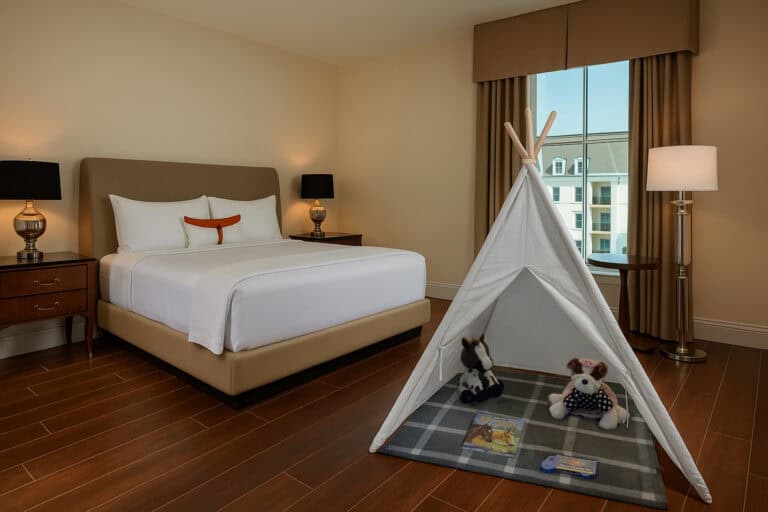 Hotel room with a bed, two nightstands, lamps, a floor lamp, and a small indoor play tent with stuffed animals and books on a rug near a window.