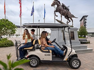 Three people ride in a golf cart past a horse statue and flagpoles in a paved outdoor area.