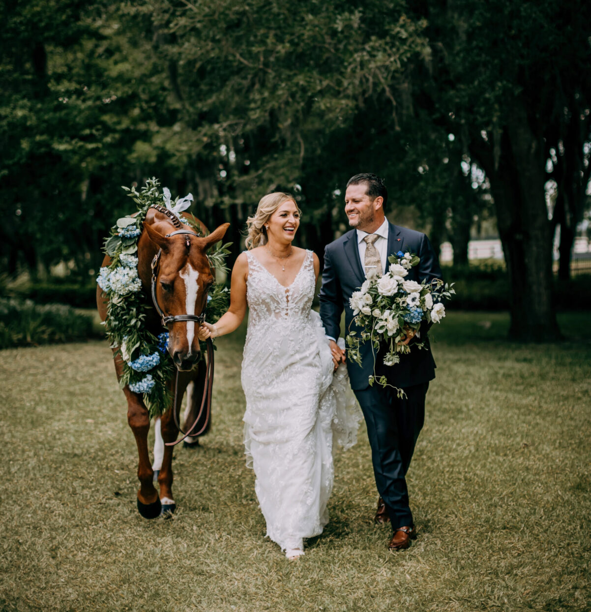 A bride and groom walk on grass, smiling and holding hands, with a brown horse decorated with greenery and flowers beside them. Tall trees fill the background.