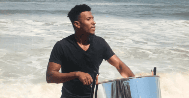 A man in a black shirt plays a steel drum by the ocean, with waves in the background and water near his legs.