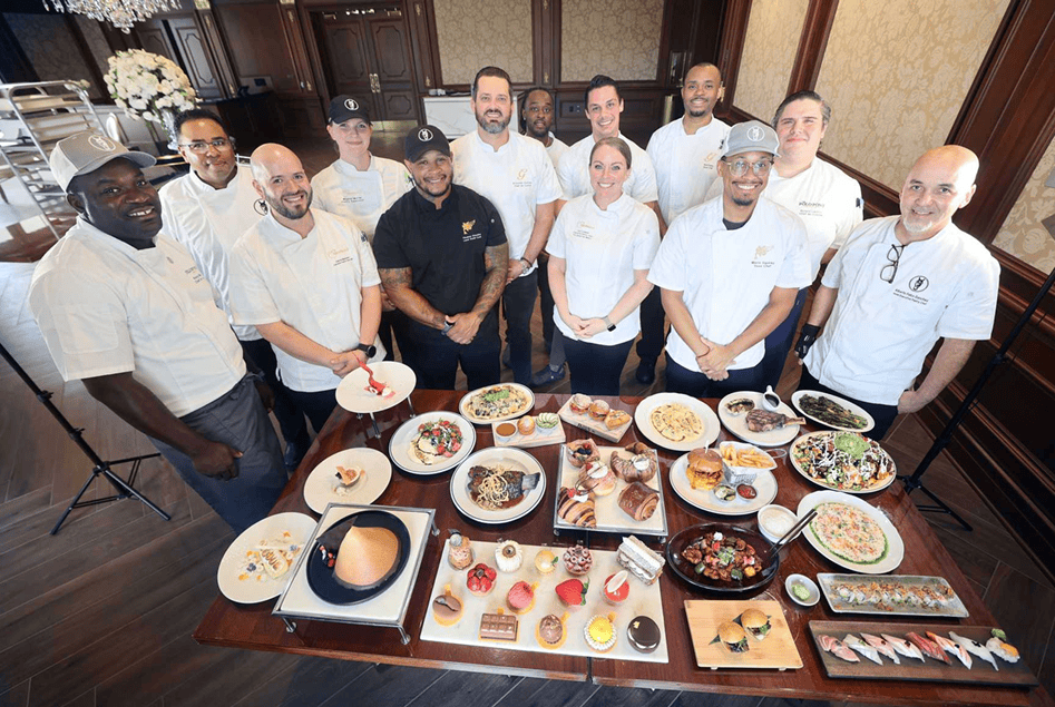 A group of chefs stands behind a large table displaying various plated dishes in an elegant dining room.