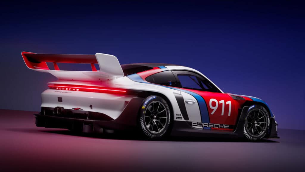 A white Porsche 911 race car with red and blue graphics is shown in a studio setting, viewed from the rear left side under dramatic lighting.