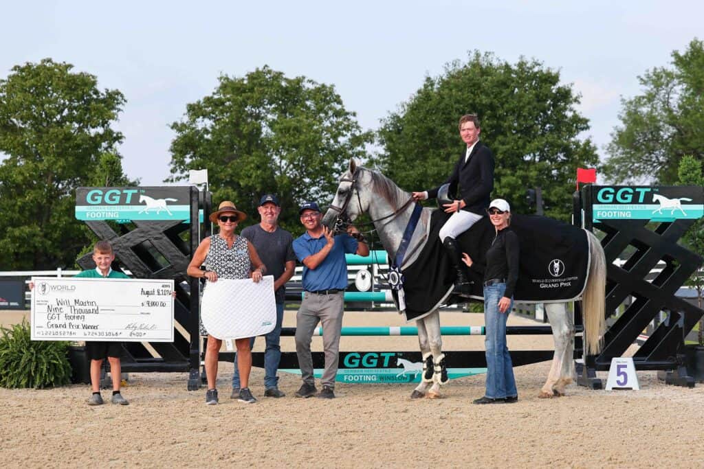 A group of people poses with a horse, rider, large check, and awards at an outdoor equestrian event, with jumps and trees in the background.