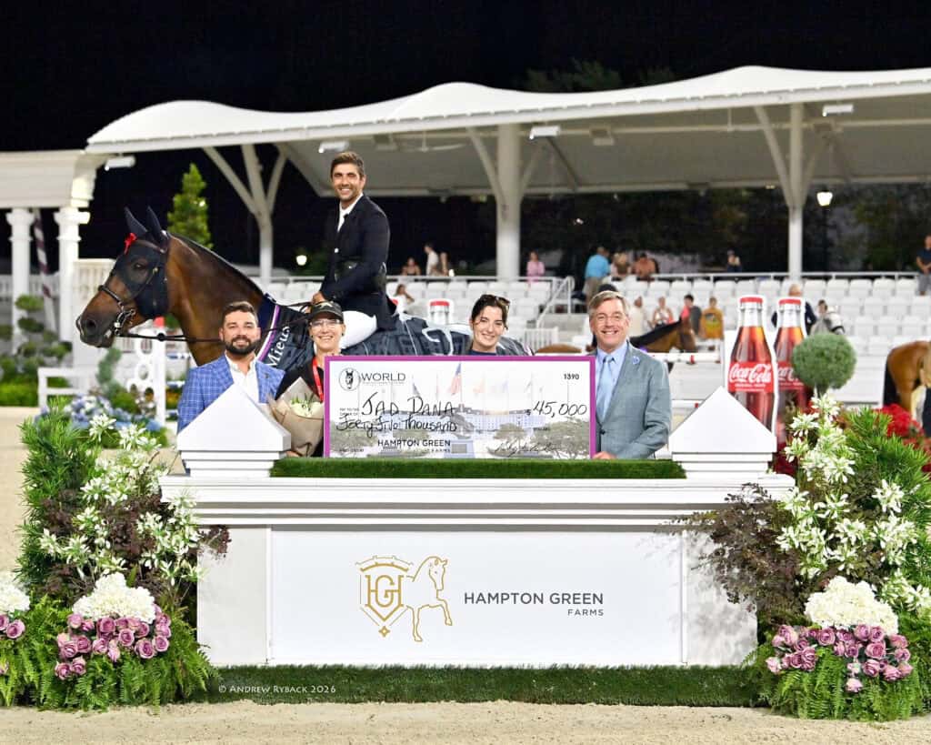 A group of people and a horse pose with a large presentation check for $145,000 at an equestrian event, with flowers and sponsor logos visible.