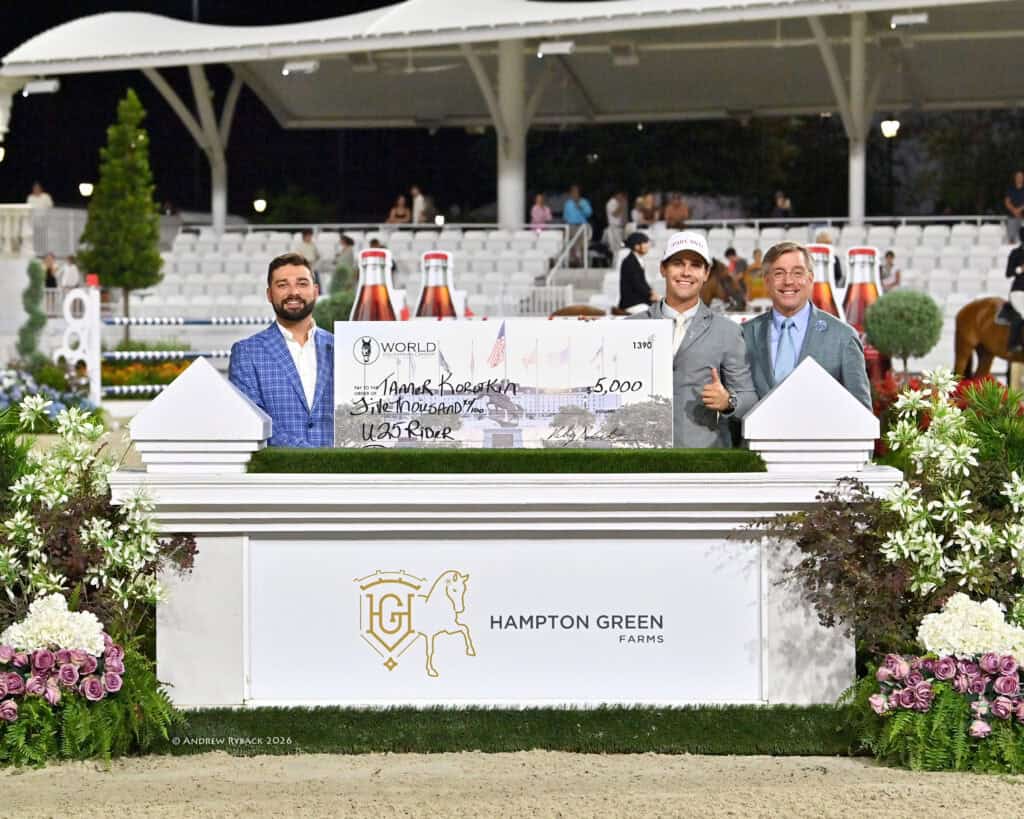 Three men stand behind a sign reading “Hampton Green Farms” holding a large $5,000 check in an outdoor arena with white seating and floral decorations.
