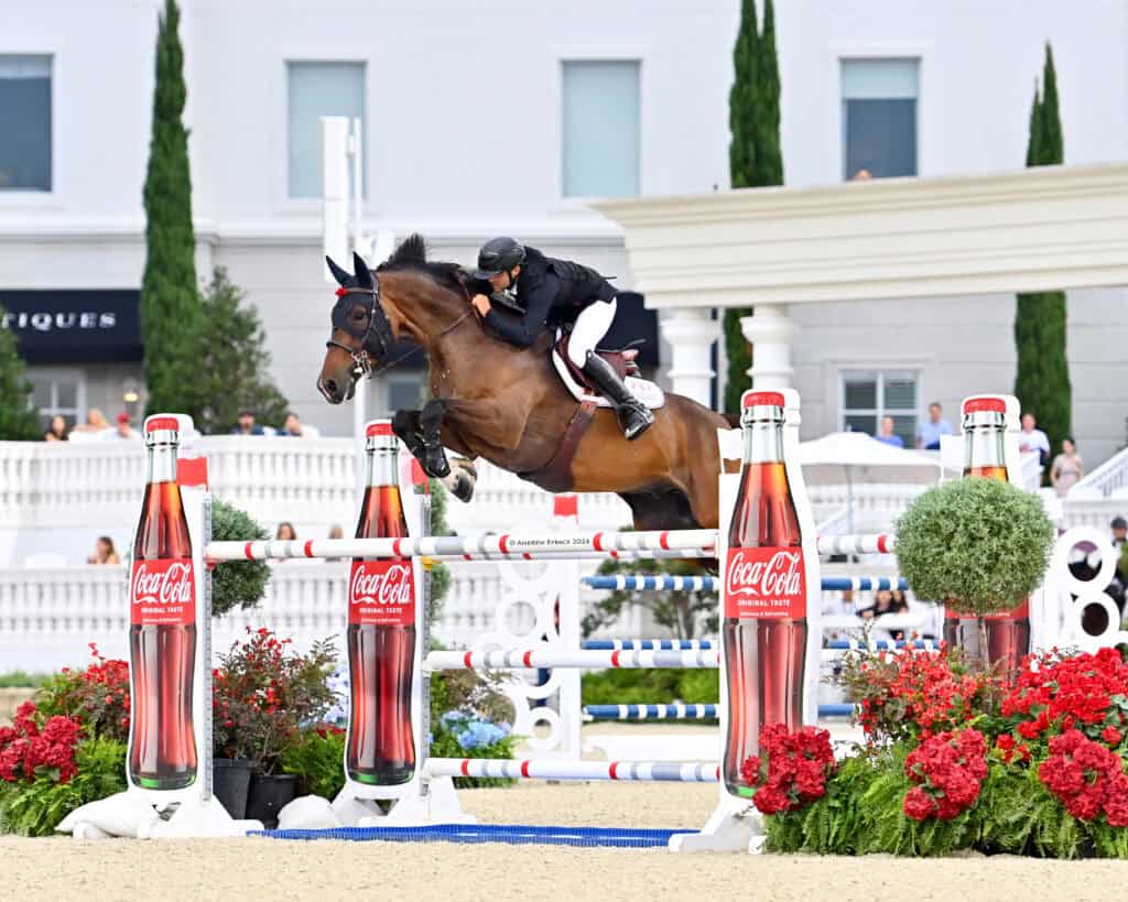 Equestrian rider on a brown horse jumps over an obstacle decorated with Coca-Cola bottles during a show jumping event.