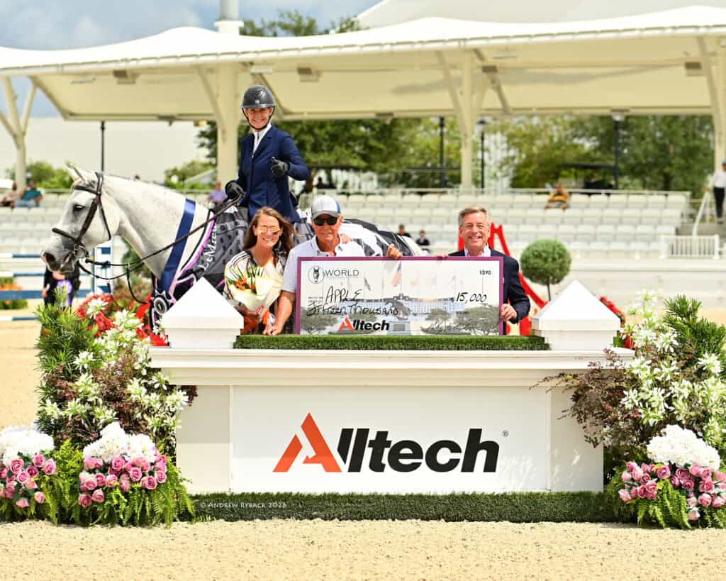 Four people pose with a horse and an oversized $15,000 check in front of an “Alltech” sign at an equestrian event.
