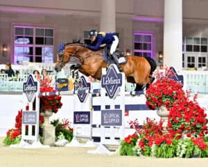 A rider wearing a helmet and jacket guides a horse over a LeMieux-branded jump decorated with red flowers during an indoor equestrian competition.