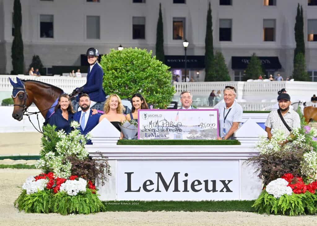 A group poses with a rider on a horse behind a large check and a LeMieux sign; the scene is outdoors with flowers and a white building in the background.