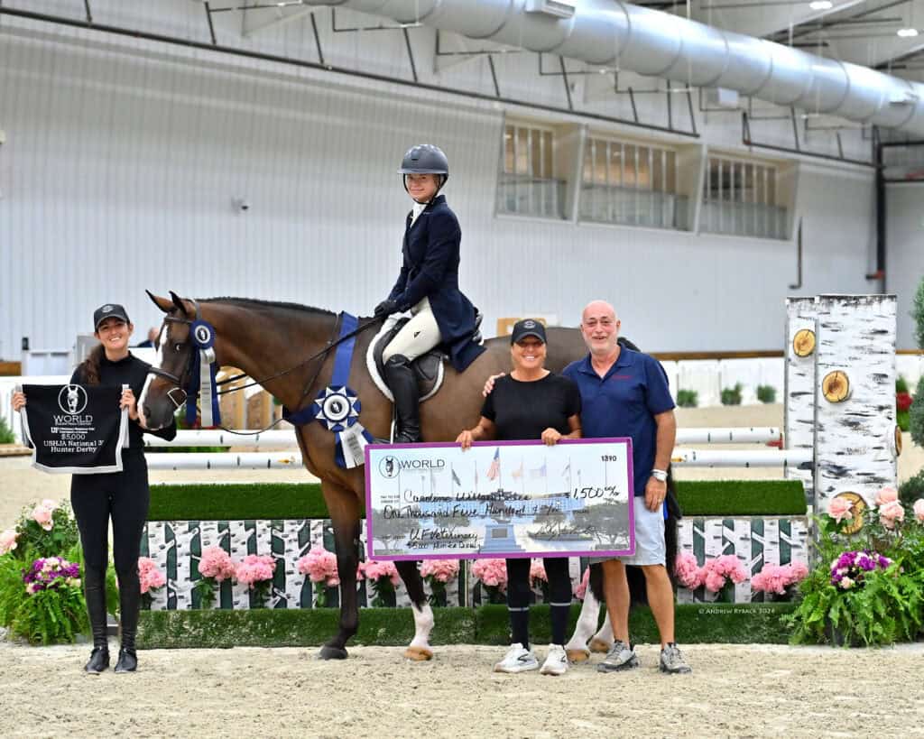 Four people and a horse pose indoors; one holds a prize sign, another sits on the horse, and two others hold a large check. A showjumping obstacle and flowers are in the background.