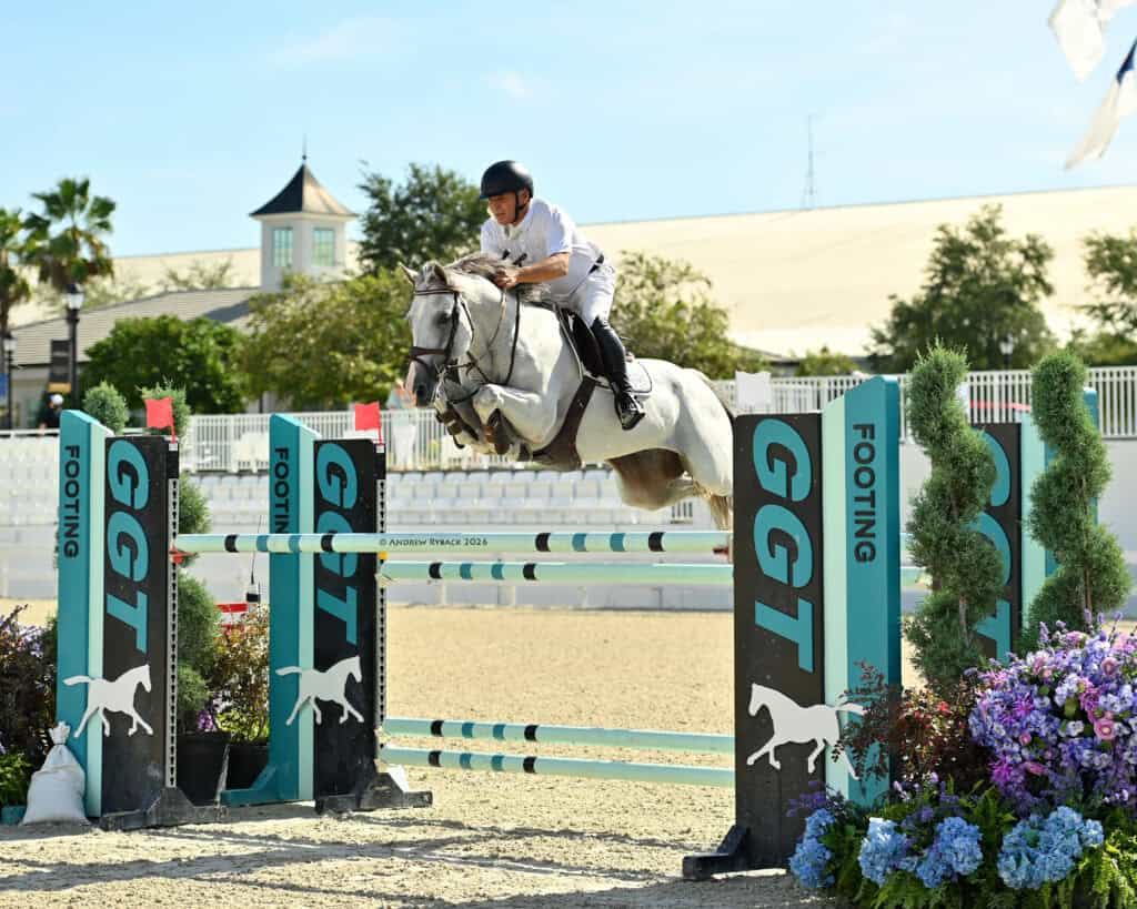 A rider in a white outfit guides a gray horse over a jump labeled “GGT Footing” in an outdoor equestrian arena on a sunny day.