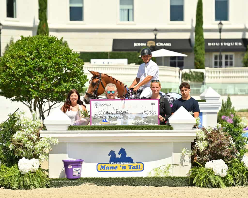 Five people, one on horseback, pose around a large check for $15,000 on a winner’s podium outdoors, in front of a building with bushes and signage.