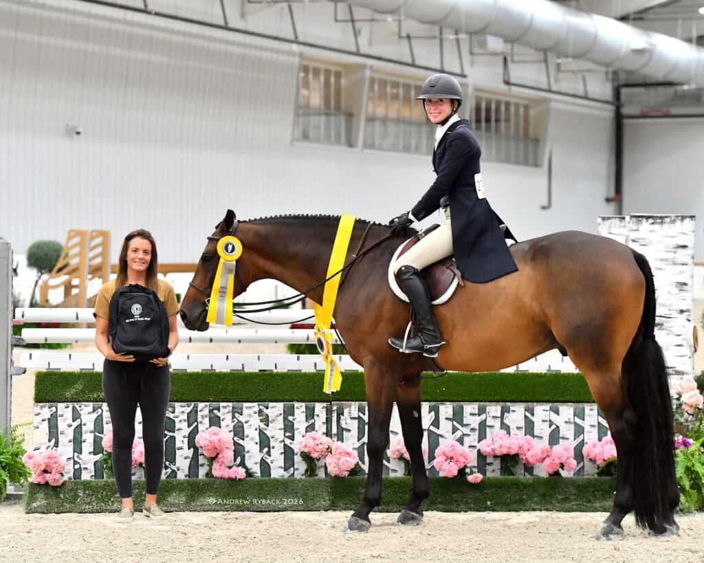 A rider on a horse with a yellow ribbon poses next to a woman holding a plaque in an indoor equestrian arena decorated with flowers.