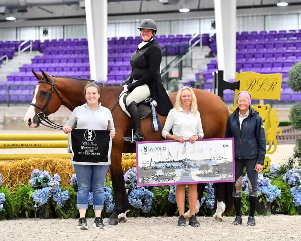 Four women pose indoors with a horse; one is mounted, while three stand holding a large check and a competition banner in front of jumping obstacles and purple stadium seating.