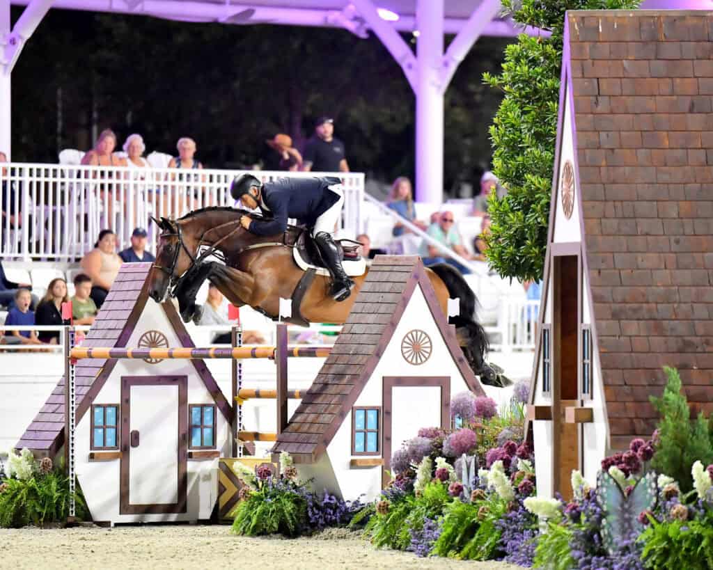 A rider on a horse jumps over a decorative obstacle designed as small houses during an equestrian event, with spectators watching from the stands.