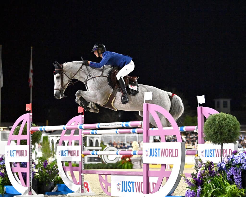 Equestrian rider in blue jacket jumps a grey horse over a pink and blue obstacle labeled “JustWorld” during a nighttime event.