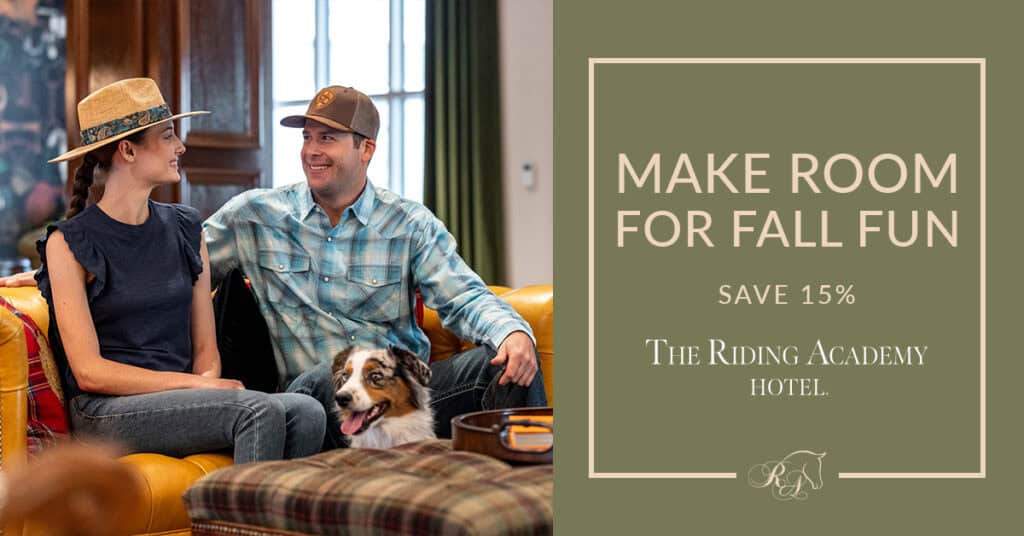 A woman and man sit smiling on a couch with a dog indoors. Text beside them offers 15% off at The Riding Academy Hotel with the phrase, “Make room for fall fun.”.