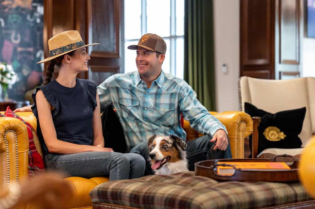 A man and woman sit on a yellow couch smiling at each other, with a dog between them and a guitar case on the table in front.