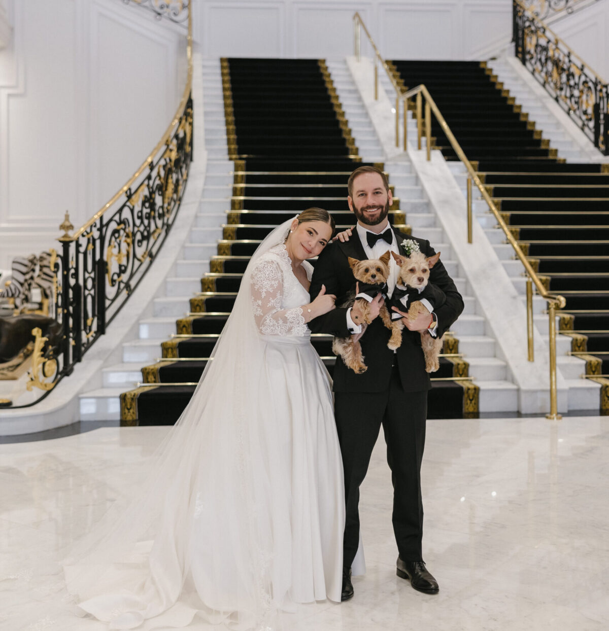 A bride and groom in formal attire stand on marble floor holding two small dogs in front of a grand staircase with gold railings and framed portraits of dogs on the wall behind them.