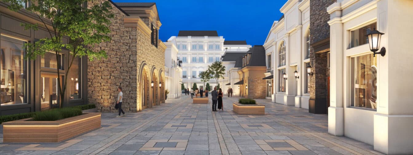 Pedestrian shopping street with stone and white buildings, large windows, benches, planters, trees, and people walking under a clear evening sky.