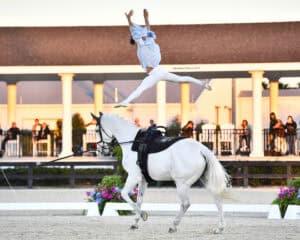 A person in mid-air performs an acrobatic leap above a white horse during an equestrian event, with spectators watching in the background.
