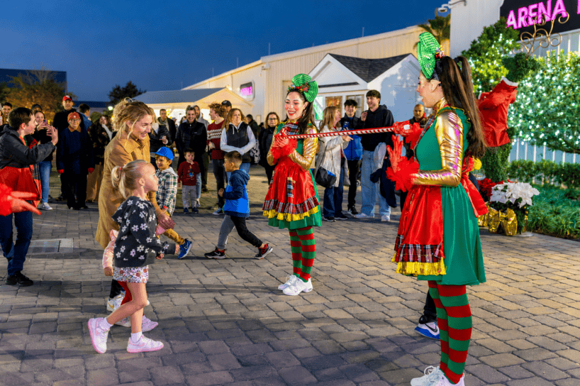 Two performers in festive costumes hold a limbo stick as children and adults, including a girl and a woman, watch or participate in the outdoor event near holiday lights and decorations at night.
