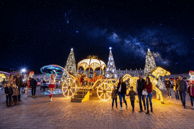 People gather around a brightly lit carriage and horse display with Christmas trees, under a starry night sky. Some stand, some take photos, and others walk by festive lights and decorations.