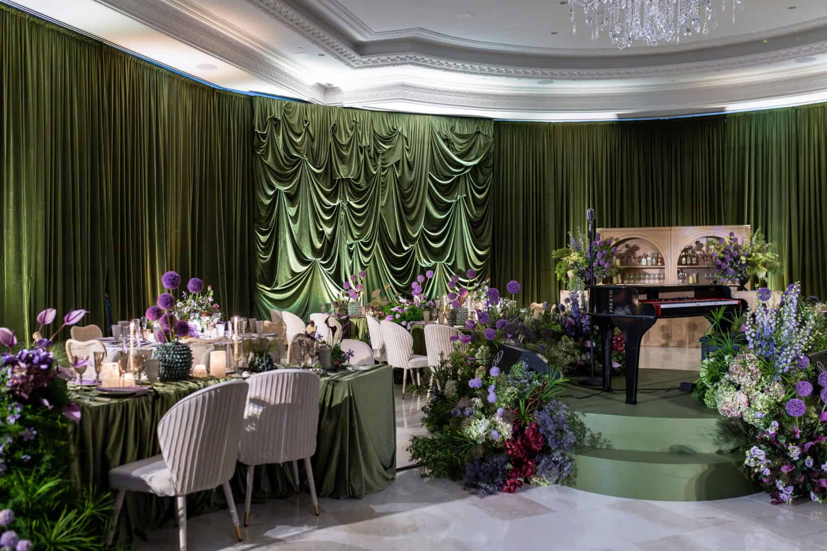 Elegant event space with green draped curtains, decorated tables, floral arrangements, a grand piano on a round stage, and a bar in the background.
