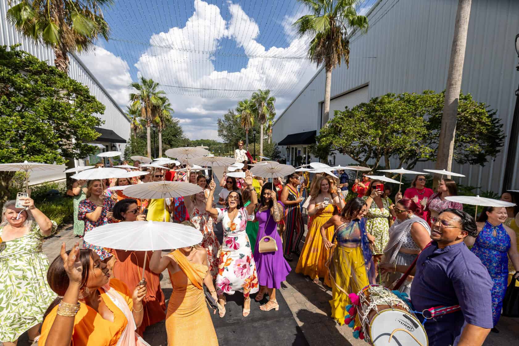 A group of people in colorful clothing stand outdoors holding white umbrellas, with palm trees and a building in the background.