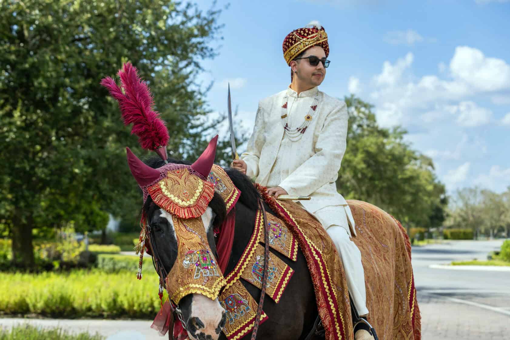 A man in traditional Indian attire and sunglasses rides a decorated horse outdoors on a sunny day.