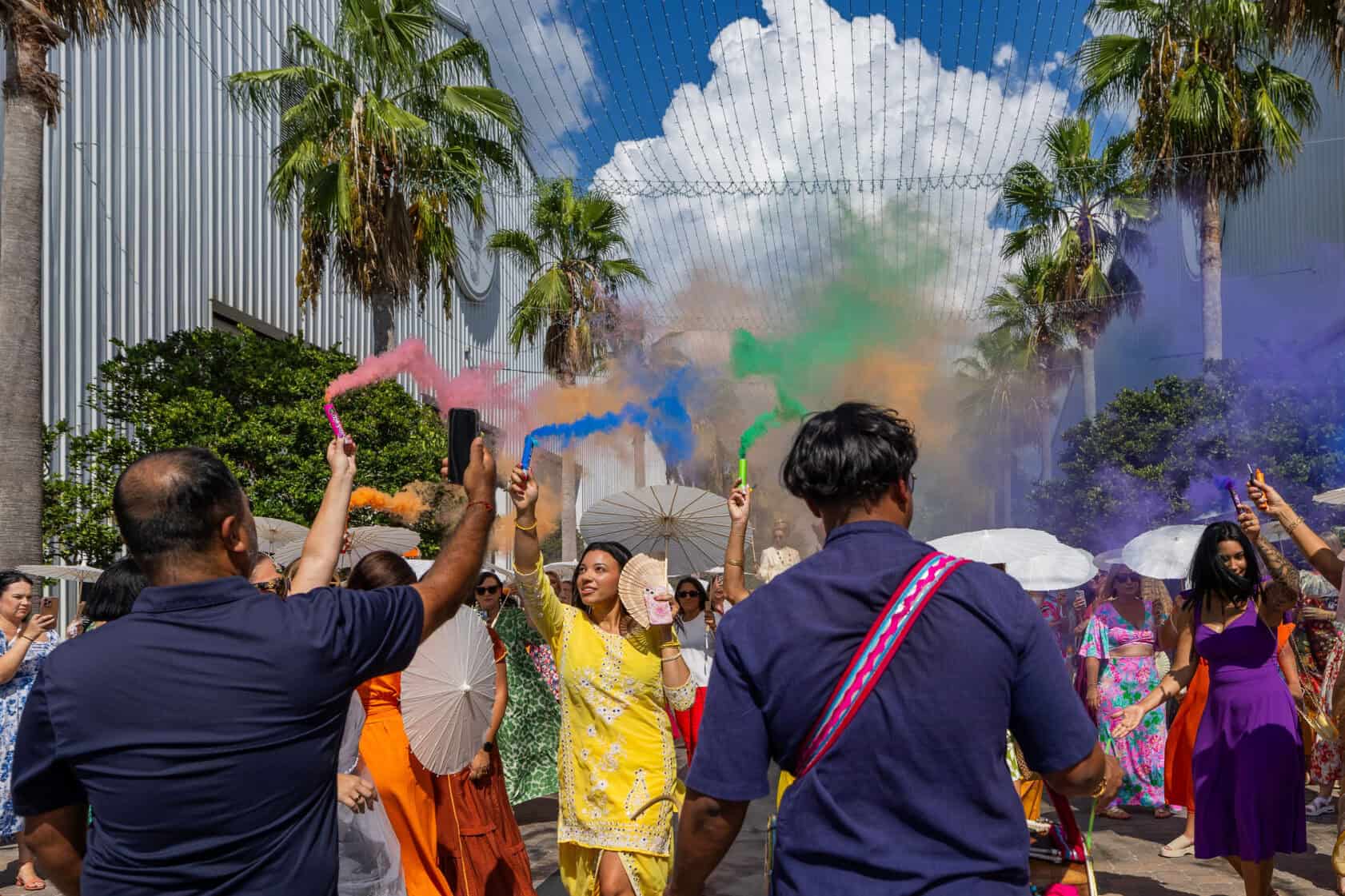 A group of people outdoors celebrate with colorful smoke, parasols, and festive clothing under palm trees and a partly cloudy sky.