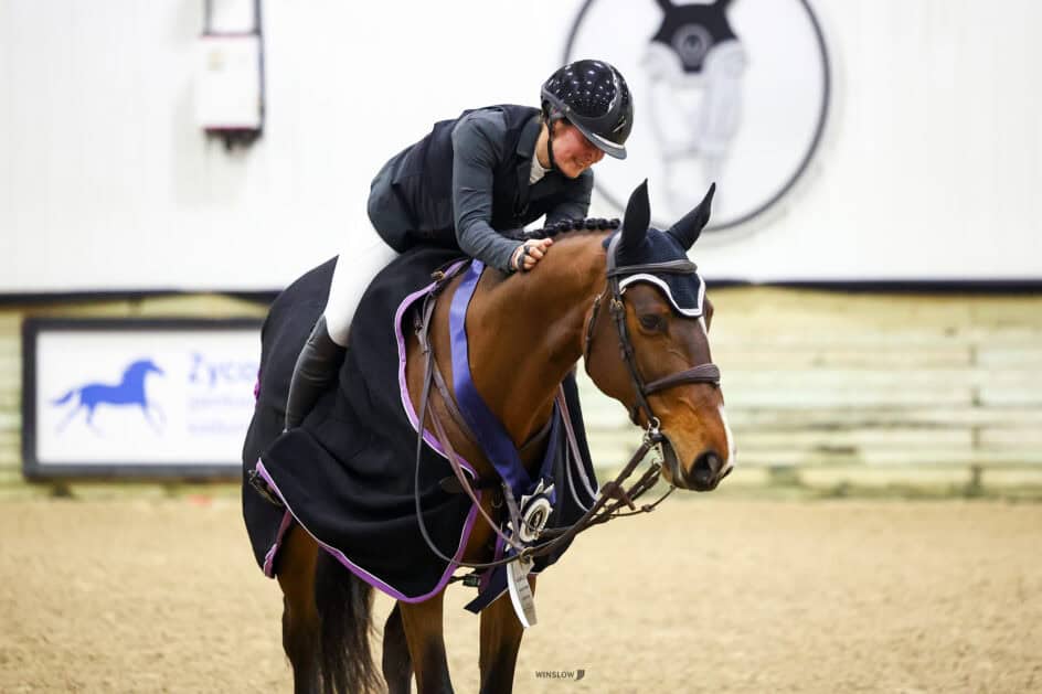 A rider wearing a helmet and jacket sits on a horse draped in a black blanket with a rosette, inside an indoor arena.
