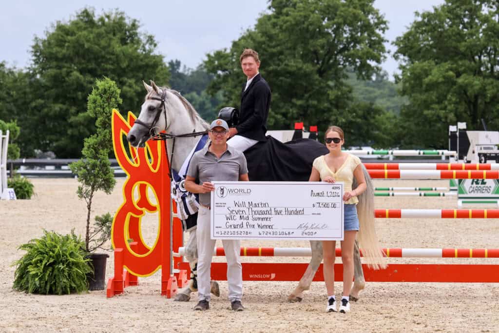 A man on horseback poses with two people holding a large prize check in front of a show jumping obstacle at an outdoor equestrian event.