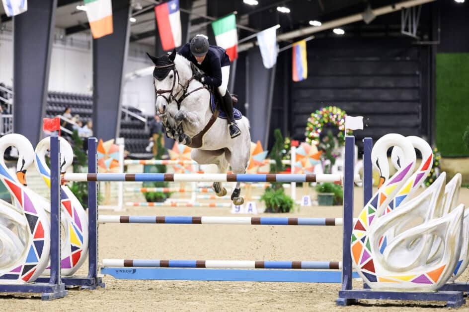 A rider on a gray horse jumps over a colorful barrier with swan designs during an indoor equestrian event.