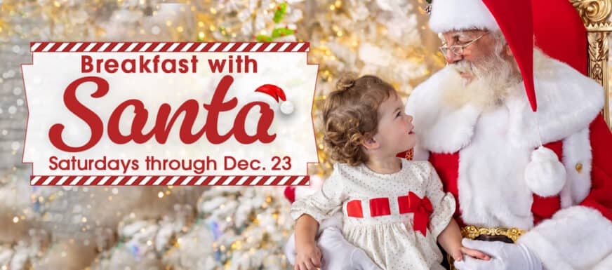 A young child in a white dress sits next to Santa Claus in front of a decorated Christmas tree. A sign reads: “Breakfast with Santa, Saturdays through Dec. 23.”.