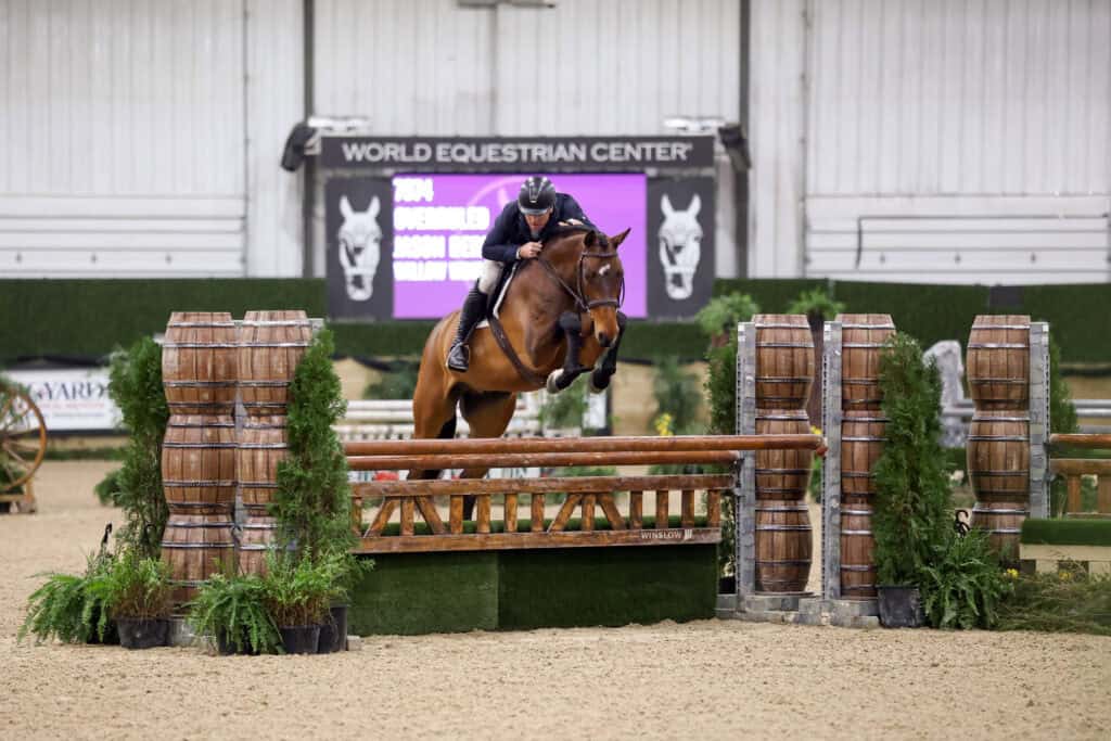 A rider on a horse jumps over a wooden fence obstacle during an indoor equestrian event at the World Equestrian Center.