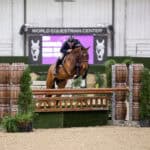 A rider on a horse jumps over a wooden fence obstacle during an indoor equestrian event at the World Equestrian Center.