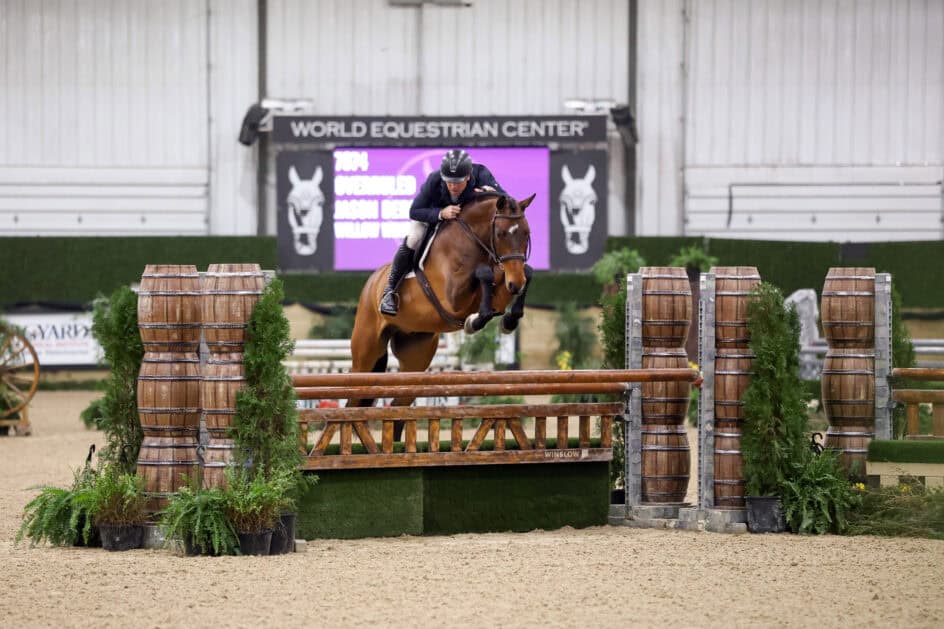 A rider on a horse jumps over a wooden fence obstacle during an indoor equestrian event at the World Equestrian Center.
