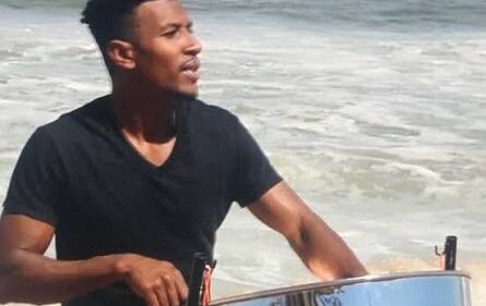 A man plays a steelpan drum on a beach with waves in the background.
