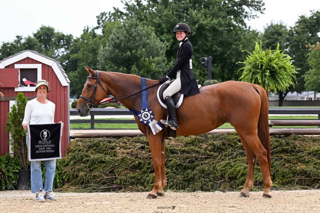 A rider in formal equestrian attire sits on a horse wearing a winner’s ribbon. A woman stands nearby holding a World Equestrian Center banner. They are outdoors with trees and a fence in the background.